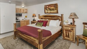 A wooden bed with a red blanket, a wooden nightstand, a wooden chair, a wooden cabinet, a painting on the wall, and a kitchen area with wooden cabinets and a microwave. - Mat-Su Resort (Wasilla)