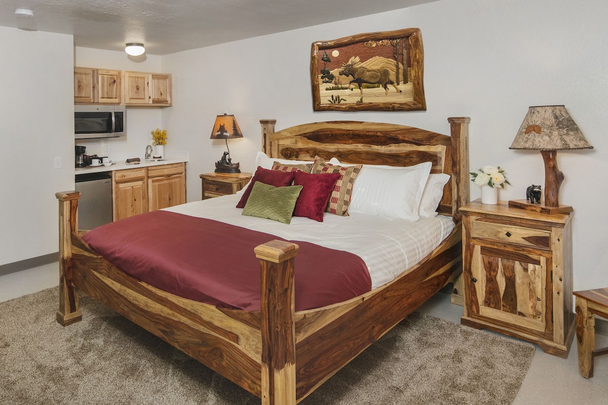 A wooden bed with a red blanket, a wooden nightstand, a wooden chair, a wooden cabinet, a painting on the wall, and a kitchen area with wooden cabinets and a microwave.