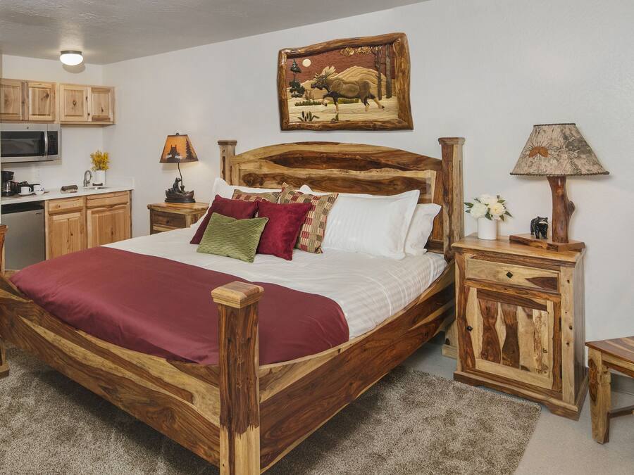 A wooden bed with a red blanket, a wooden nightstand, a wooden chair, a wooden cabinet, a painting on the wall, and a kitchen area with wooden cabinets and a microwave.