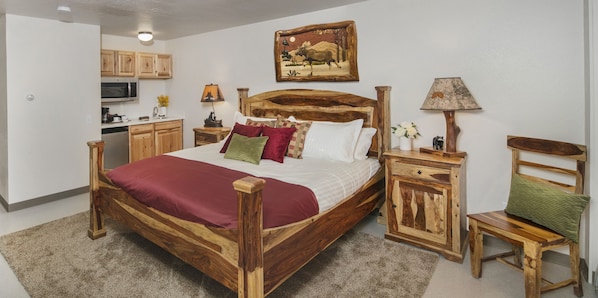 Una cama de madera con una manta roja, una mesita de noche de madera, una silla de madera, un armario de madera, un cuadro en la pared y una zona de cocina con armarios de madera y un microondas.