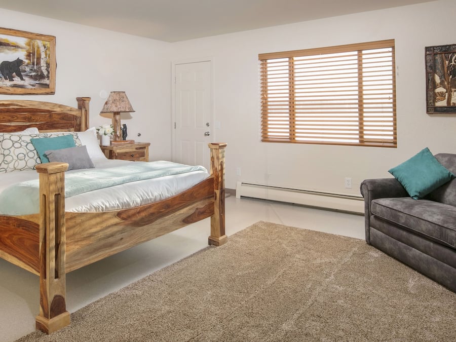Un dormitorio con cama de madera, dos mesitas de noche, un sofá gris y una ventana con persianas.