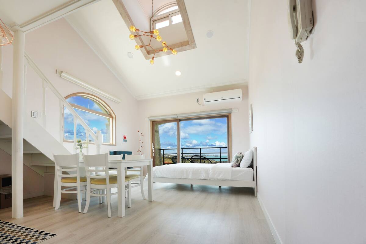 A spacious room with a bed, a dining table with chairs, a staircase, and large windows offering a view of the sea.