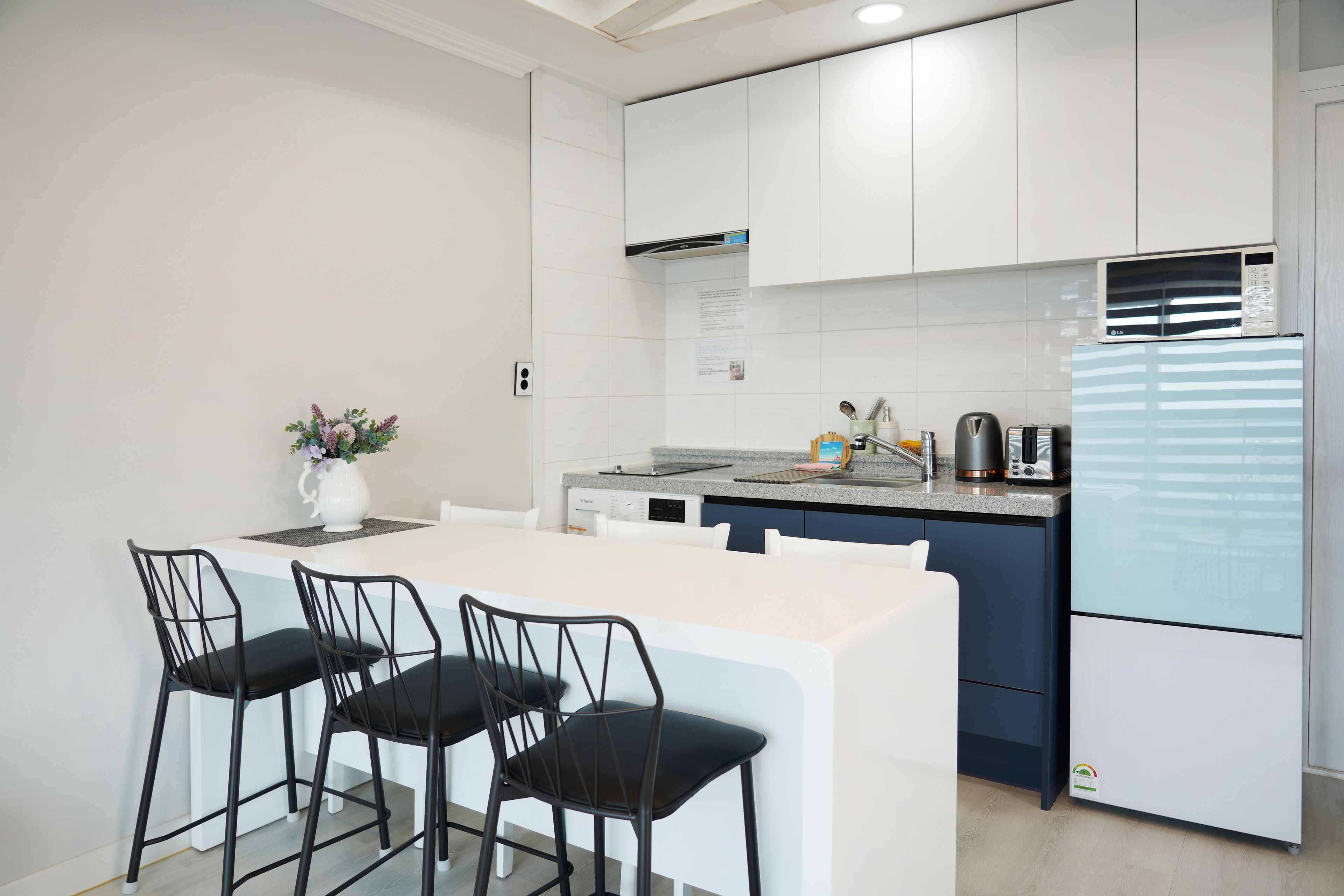 A modern kitchen with white cabinetry, a built-in microwave, and a refrigerator. There are three black chairs around a white island.