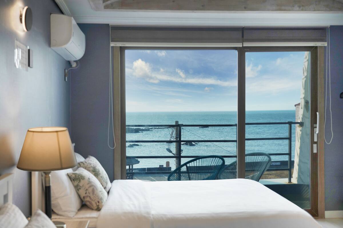 A hotel room with a bed, a lamp, a view of the sea, and a balcony with chairs.