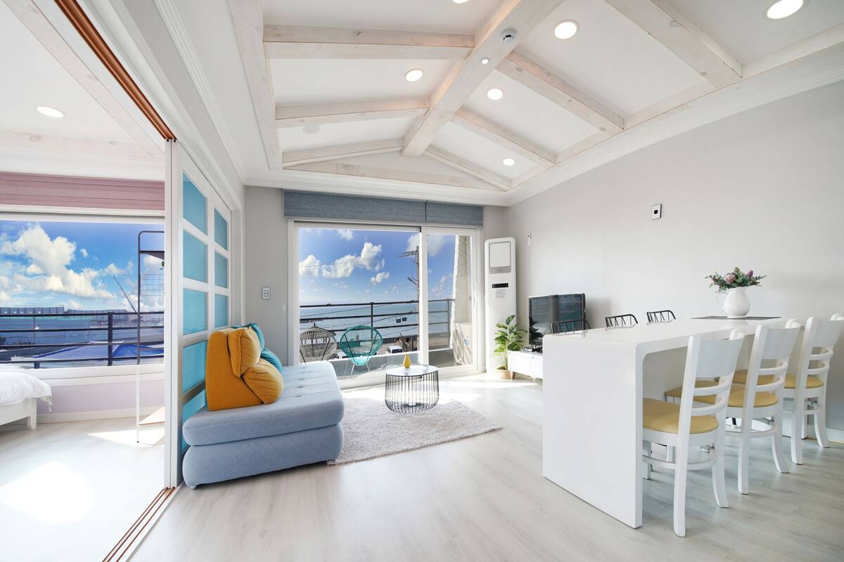 A modern living room with a sofa, a dining area with chairs, a TV, and a view of the sea.
