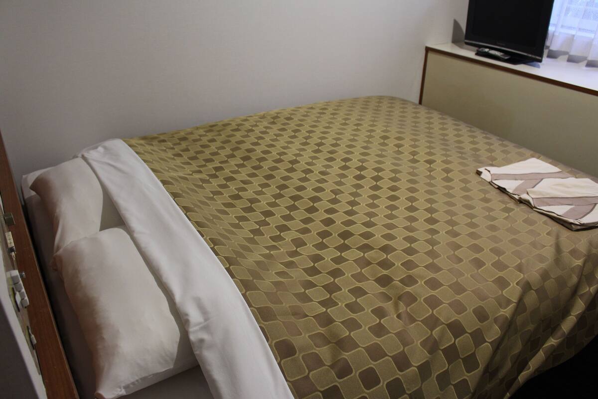 A single bed with a patterned bedspread, a pillow, and a remote control on a bedside table.