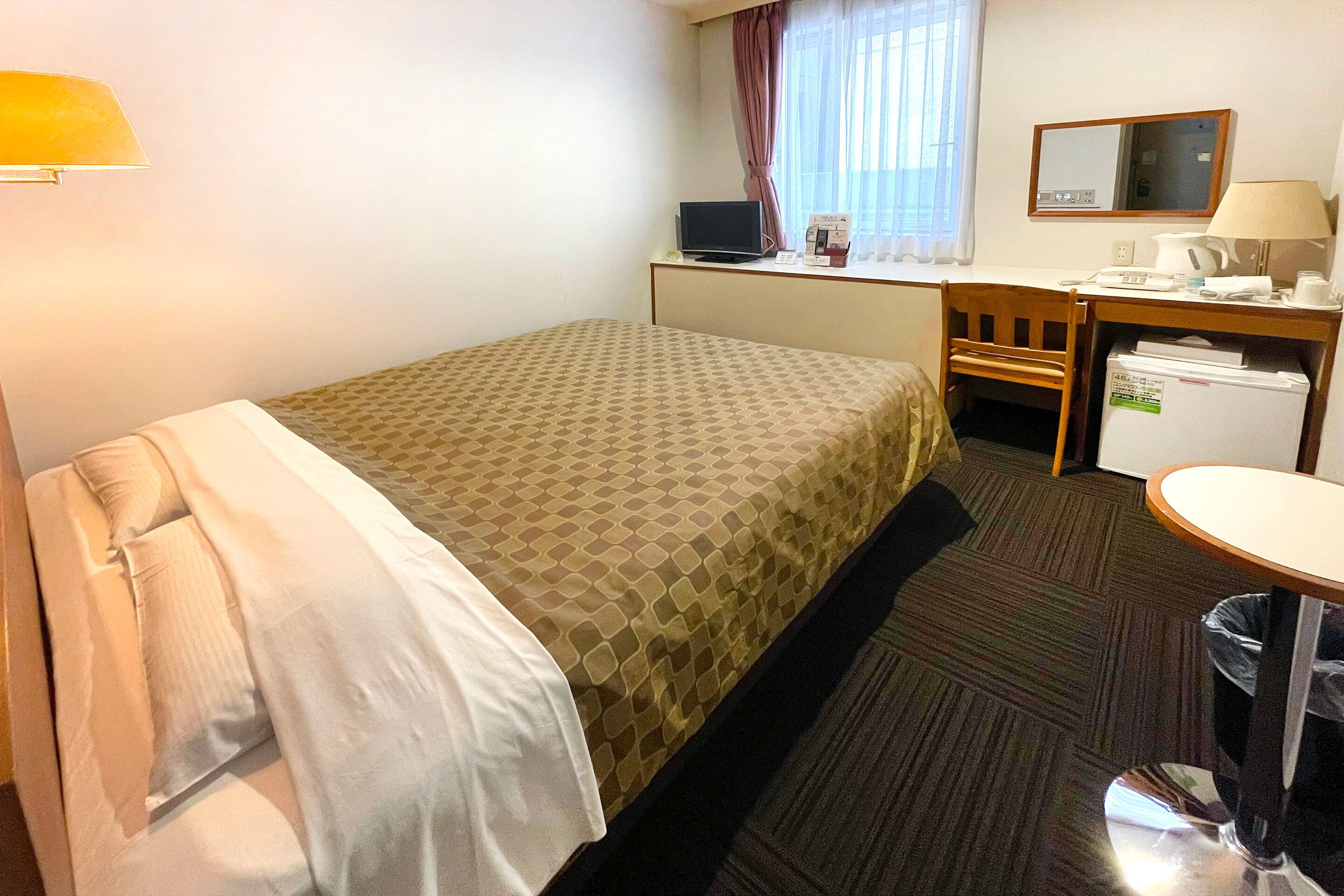 A hotel room with a bed, a desk, a chair, a small refrigerator, a lamp, and a window with curtains.