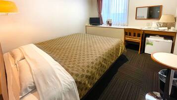 A hotel room with a bed, a desk, a chair, a small refrigerator, a lamp, and a window with curtains.