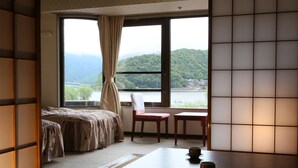 Una habitación con una cama, una mesa con tazas, una silla y vistas a un río y montañas a través de una ventana.