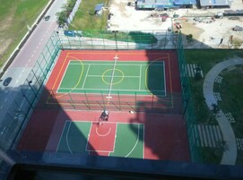 Vista aérea de una cancha polideportiva con zonas para baloncesto y fútbol, rodeada por una valla verde y cerca de una carretera.