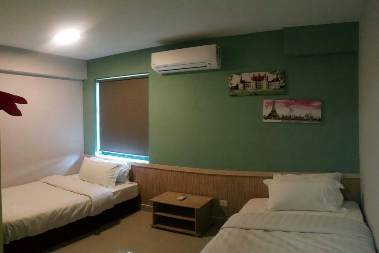 A room with two single beds, a small wooden nightstand, a window with blinds, and an air conditioning unit mounted on the wall.