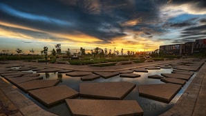 Une place publique agrémentée d’éléments aquatiques aux formes géométriques, avec un coucher de soleil en arrière-plan.
