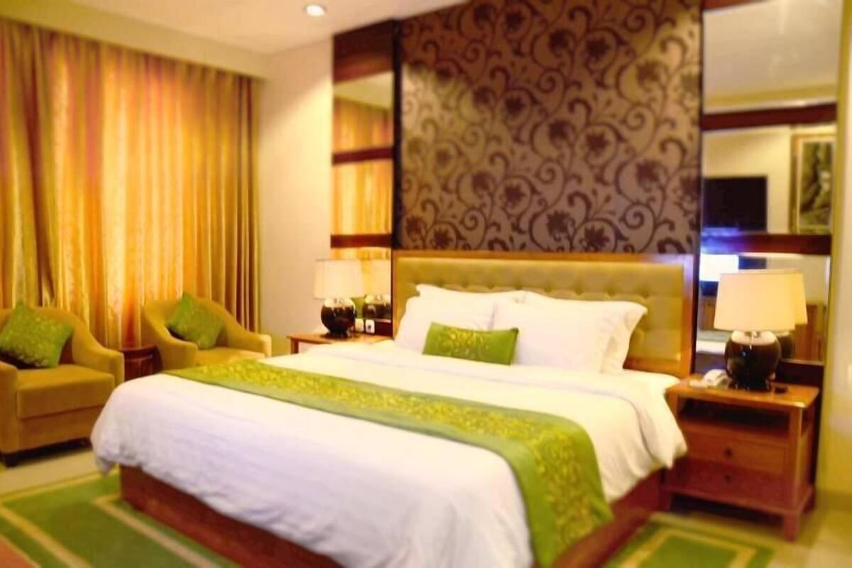 A hotel room with a large bed, two armchairs, a nightstand with a lamp, and a patterned wall.