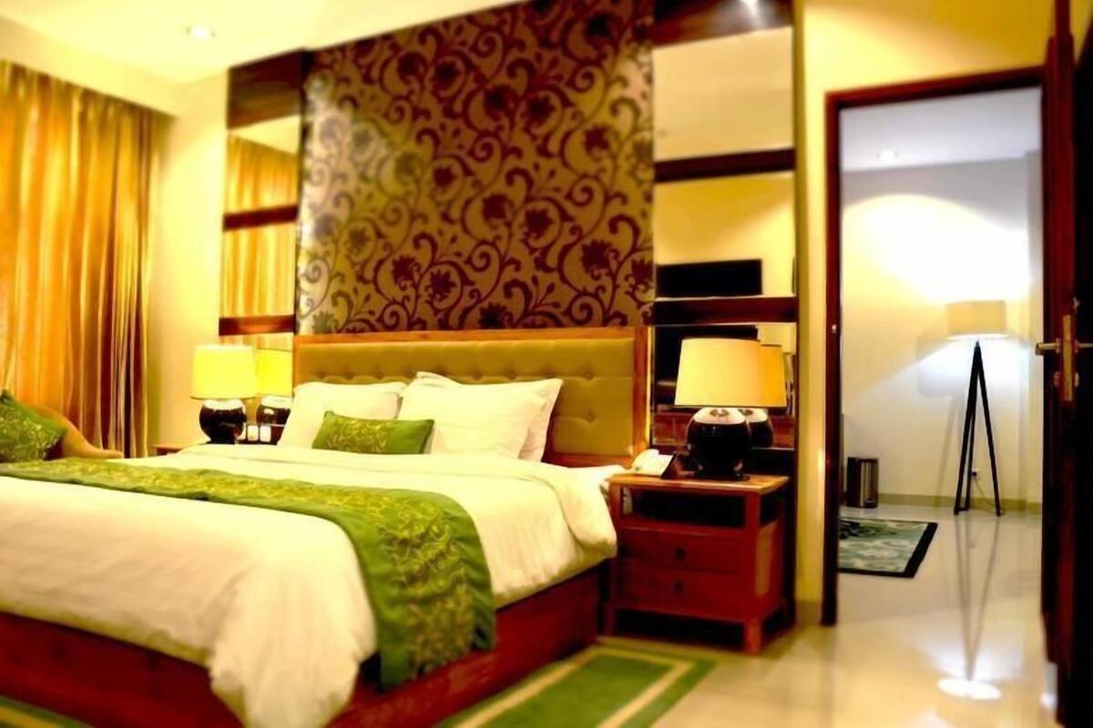 A hotel room with a large bed, two bedside tables with lamps, a patterned wallpaper, and a door leading to another room.