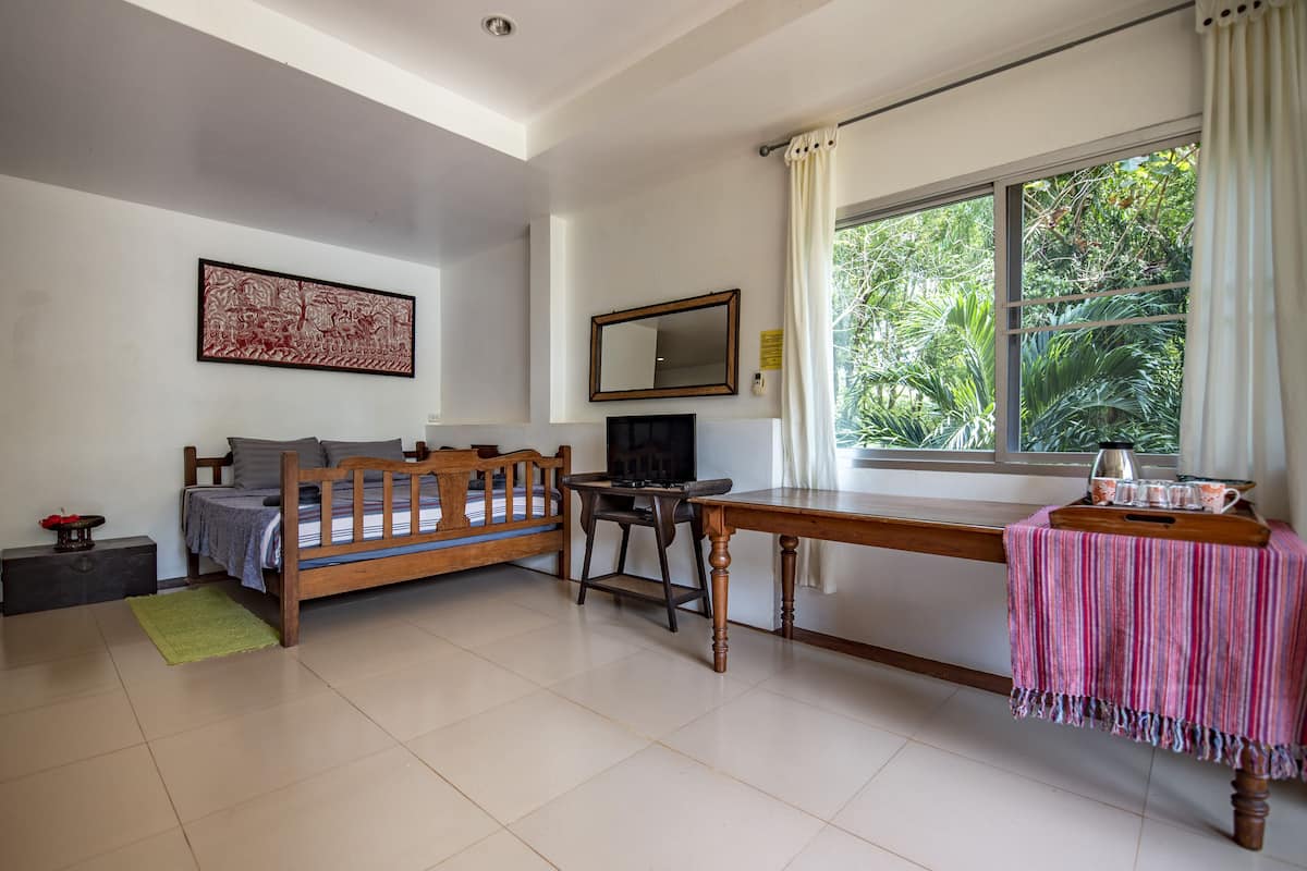 A room with a bed, a desk, a chair, a TV, and a window with a view of greenery.