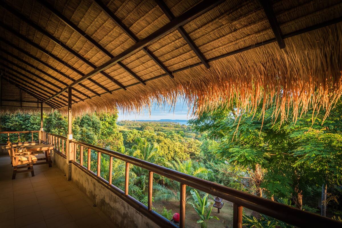 A covered outdoor area with a thatched roof, wooden benches, and a view of a lush landscape.