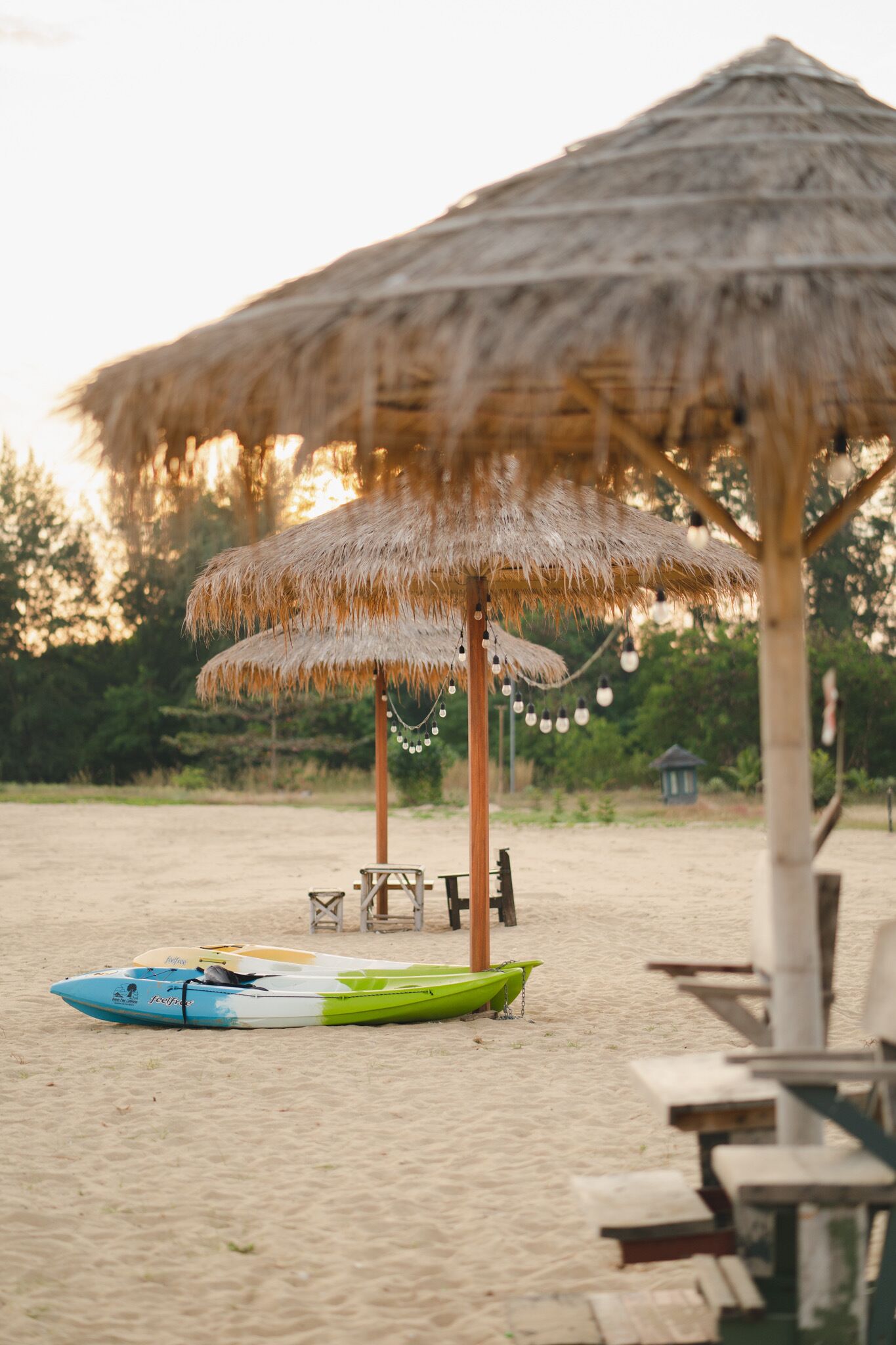 A beach with a thatched umbrella, a surfboard, and a small wooden structure in the background.