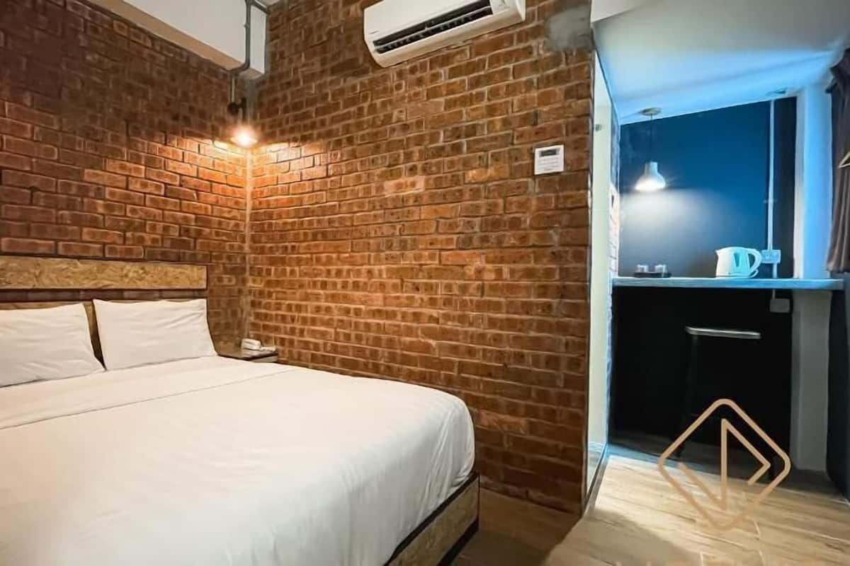 A room with a brick wall, a bed with white bedding, an air conditioning unit, and a small kitchenette area.