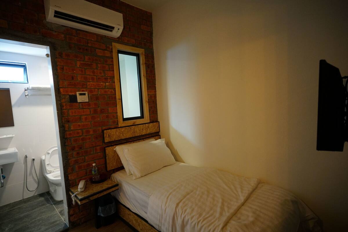 A small, compact room with a bed, a window, a brick wall, a white sink, and a toilet.