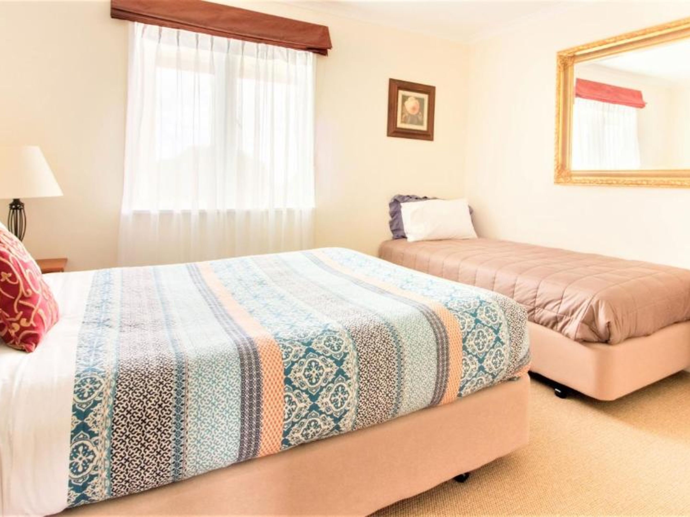 Une chambre avec un grand lit, un plus petit lit, une fenêtre avec des rideaux, une table de chevet avec une lampe et un tableau encadré au mur.