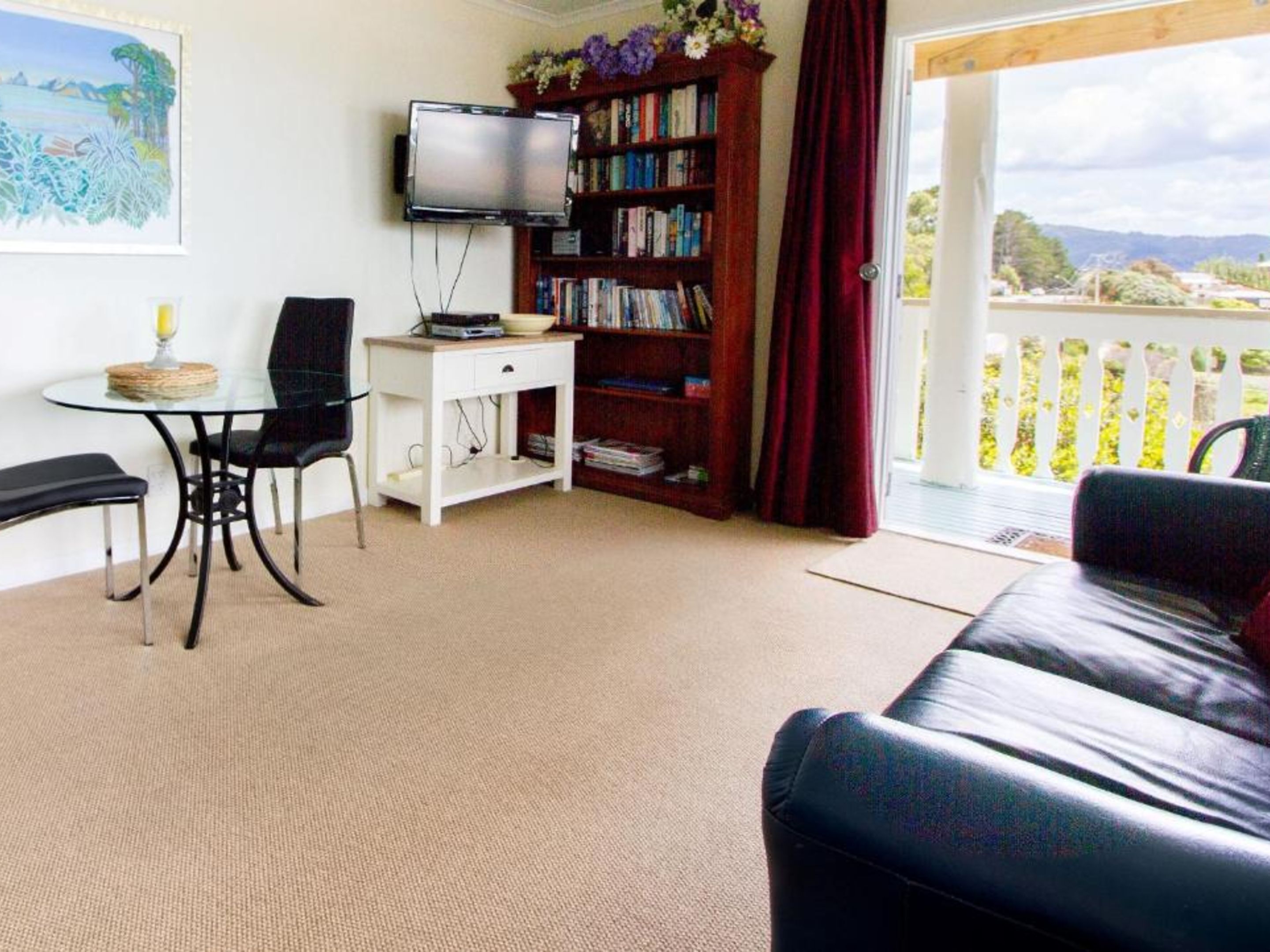Ein gemütliches Wohnzimmer mit Ledersofa, einem kleinen Tisch mit Stuhl, einem Bücherregal, einem Fernseher und einem Balkon mit Landschaftsblick.