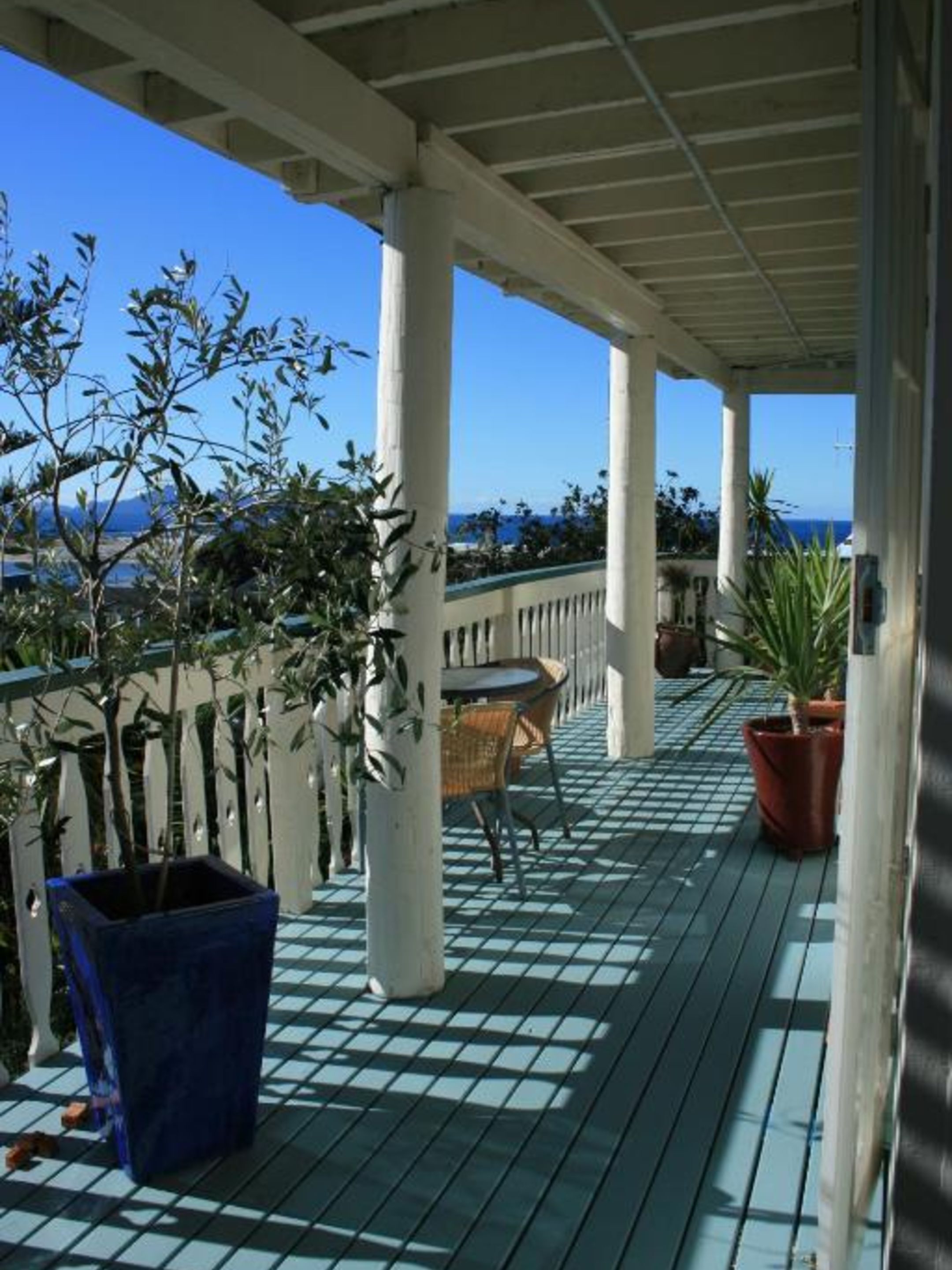 A covered outdoor area with a view of the ocean, featuring a table and chairs, potted plants, and a tiled floor.