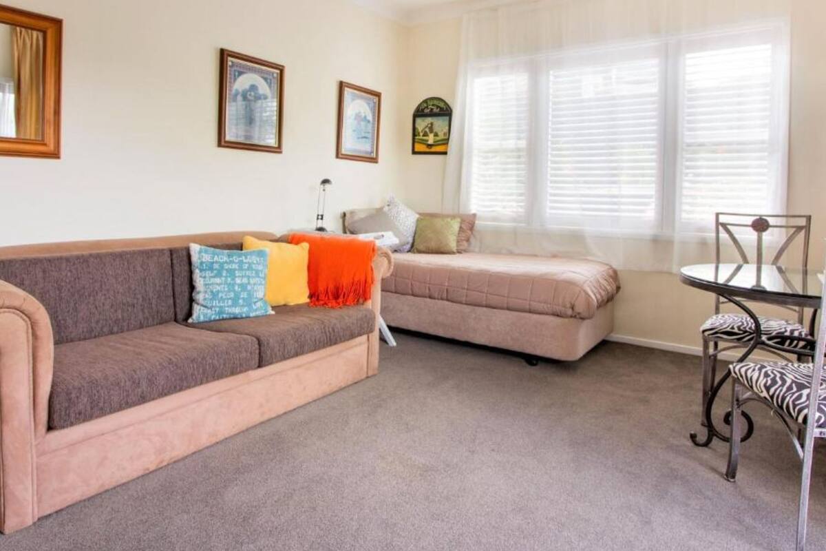 Ein gemütliches Wohnzimmer mit einer Couch, einem kleinen Sofa, einem Tisch und einem Sessel. An den Wänden hängen Bilder und es gibt ein Fenster mit Jalousien.