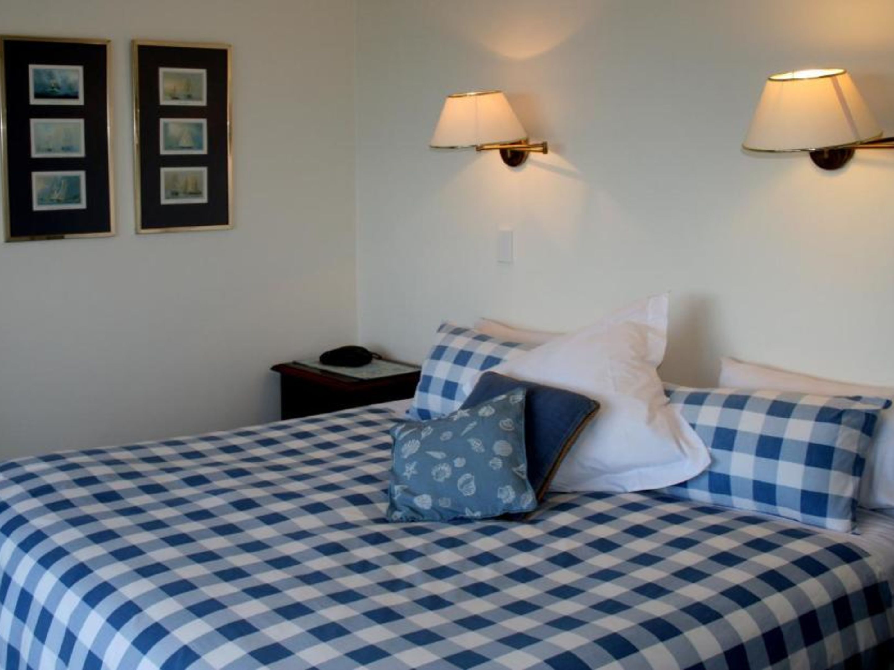 Una camera d'albergo con una coperta a quadri, due lampade fissate al muro e immagini incorniciate.