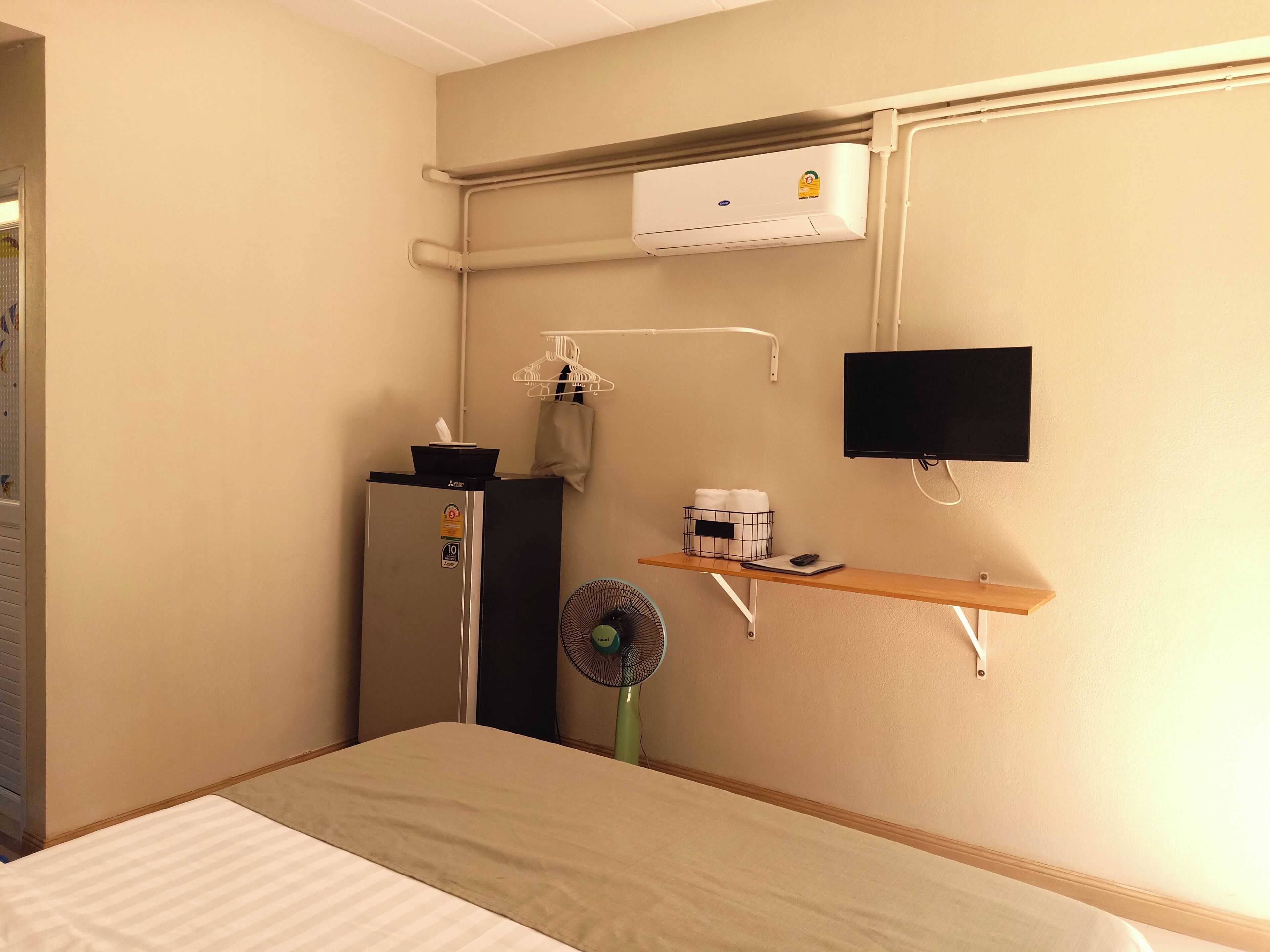 A hotel room with a bed, a small desk with a fan, a TV, an air conditioning unit, and a shelf with toiletries.