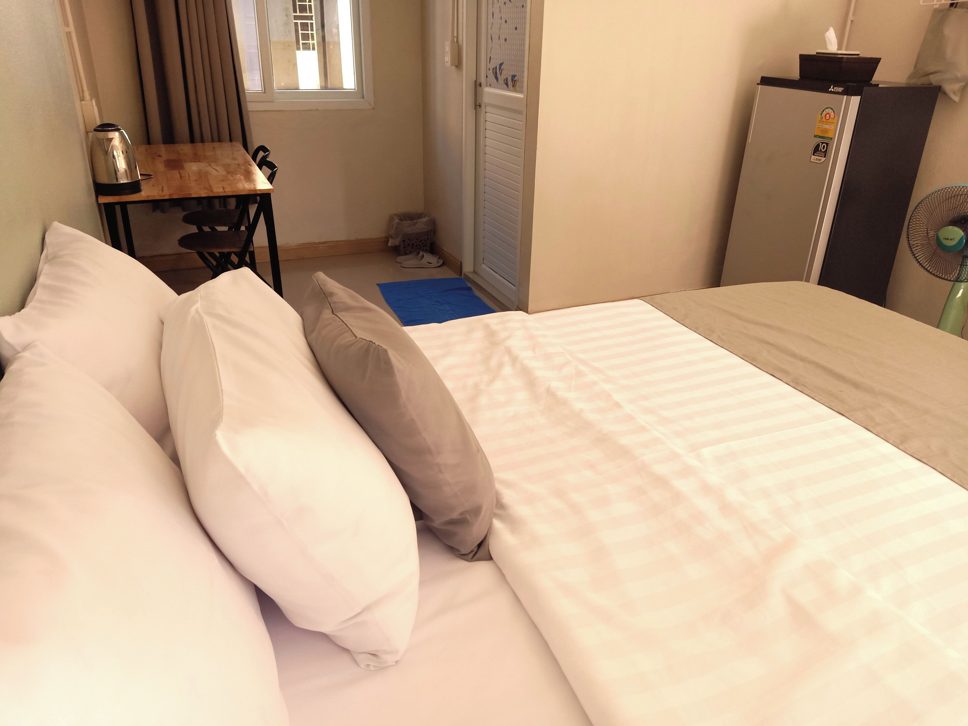 A hotel room with a bed, pillows, a desk, a chair, a radiator, a fan, and a window.