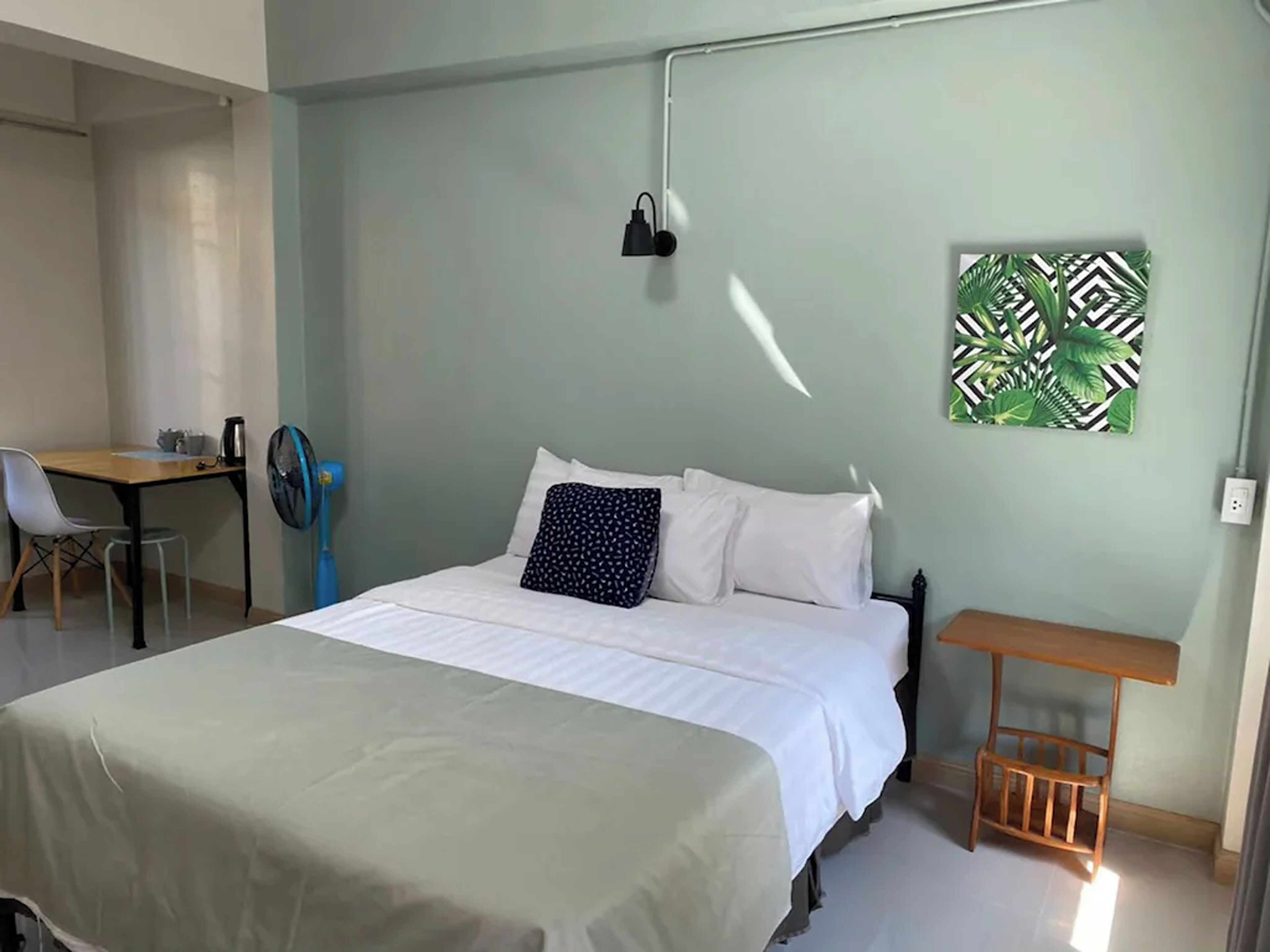 Un dormitorio con cama, escritorio, silla, una mesita y un ventilador de pared.