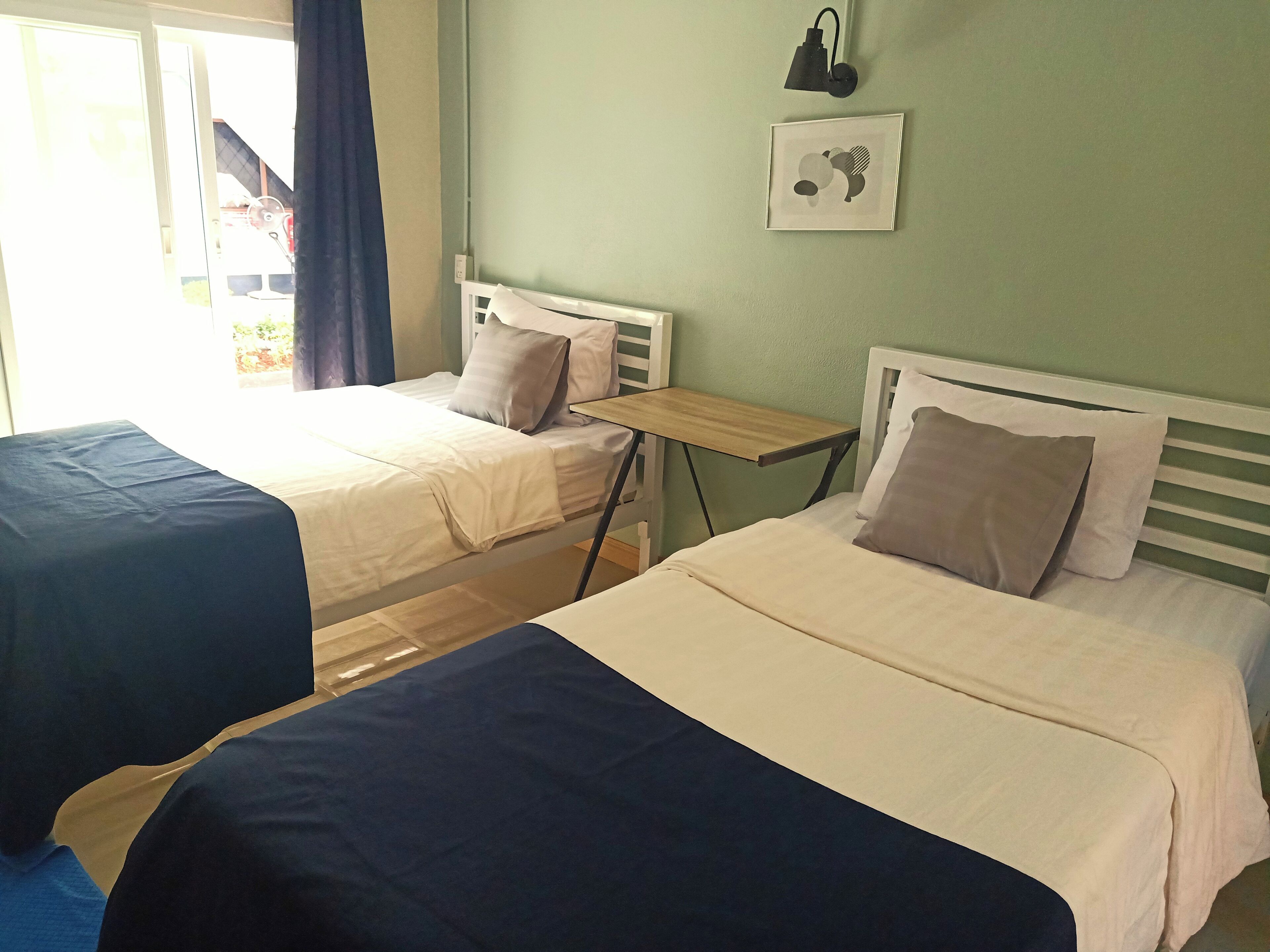 Dos camas individuales con ropa de cama blanca y azul, una mesita de madera, una lámpara de pared y un cuadro enmarcado.