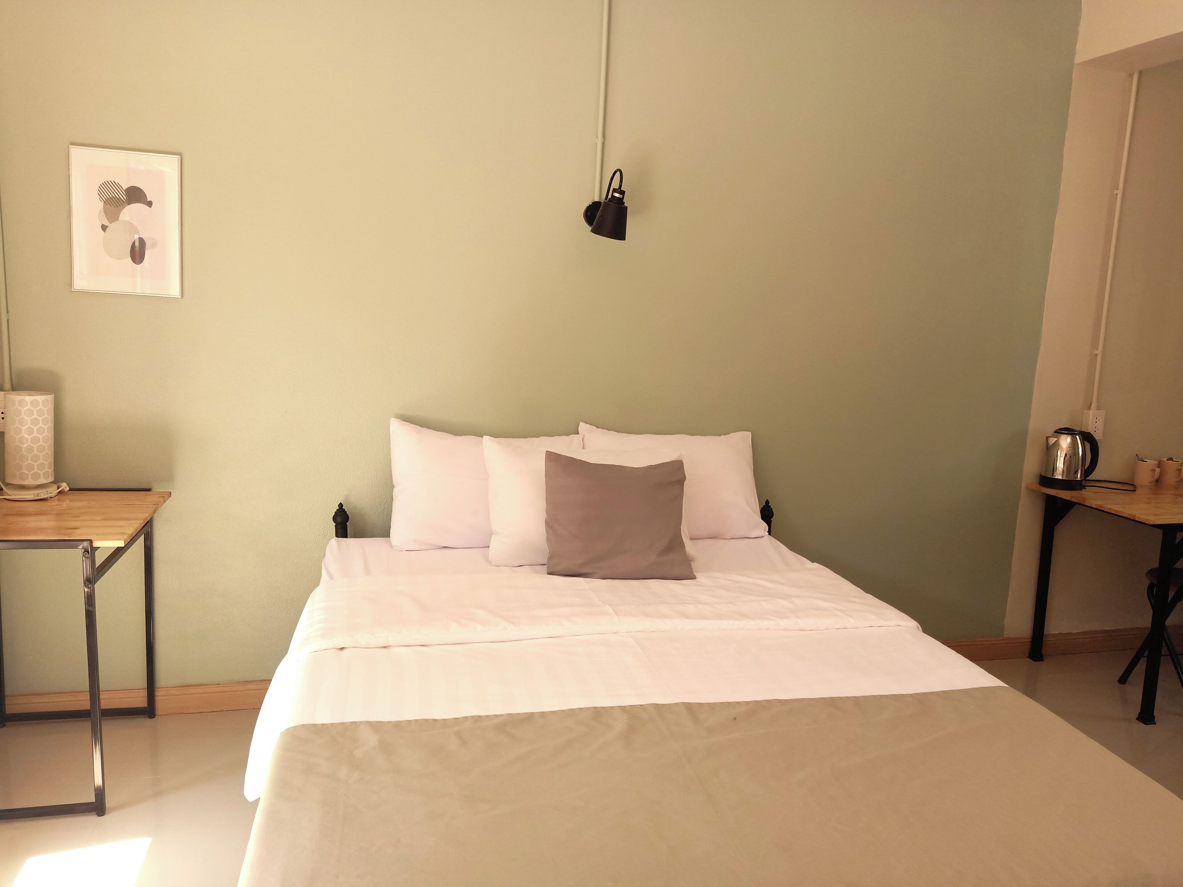 A neatly made bed with white and beige bedding, a small bedside table with a lamp, and a wall-mounted light fixture.
