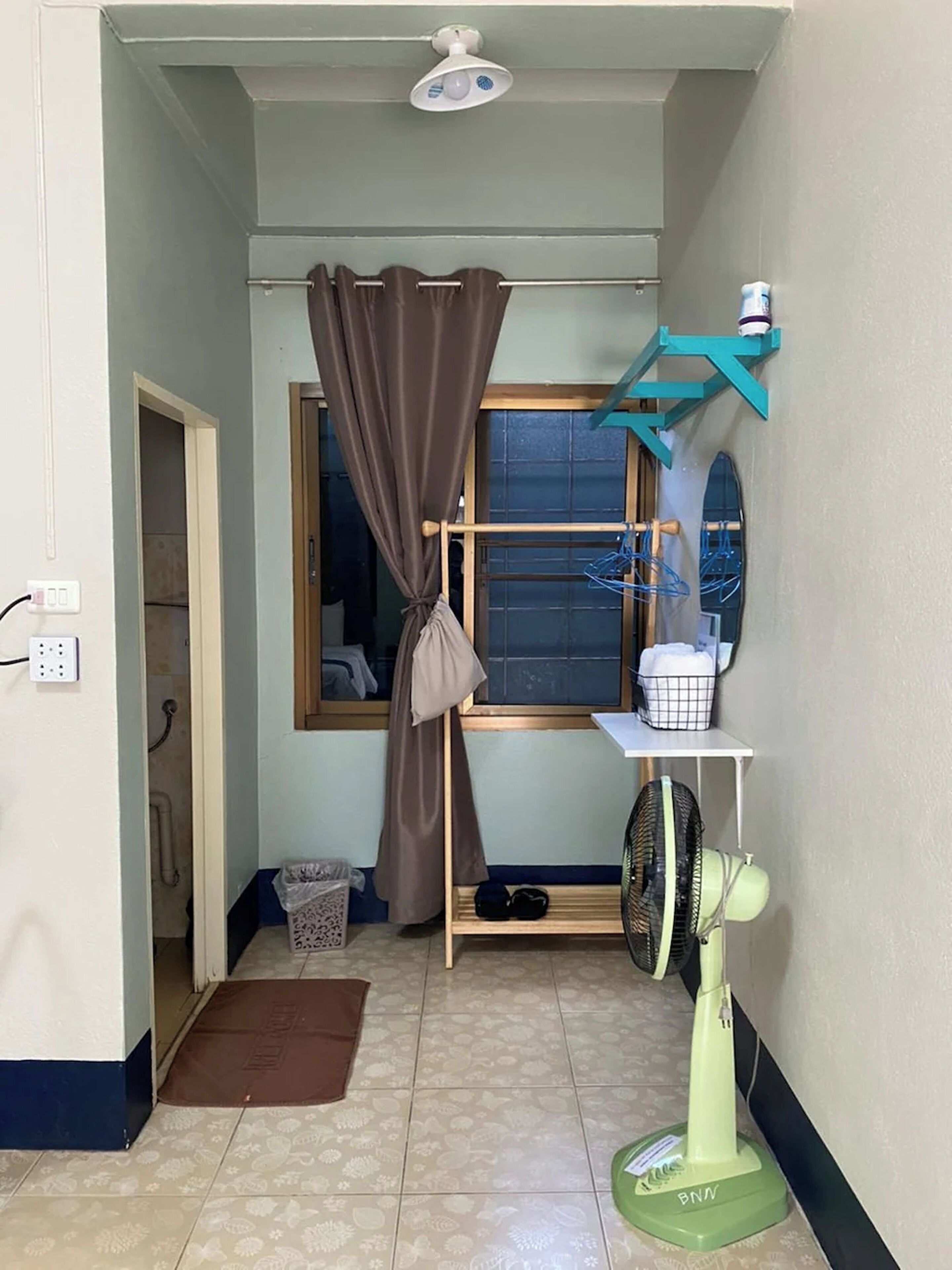 A small, narrow room with a window, a ceiling fan, a shelf with a fan, a drying rack, and a door leading to another room.