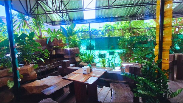 An indoor garden with a wooden table and benches, surrounded by various plants and a rock waterfall. - Rayong Lanna Hotel (Rayong)