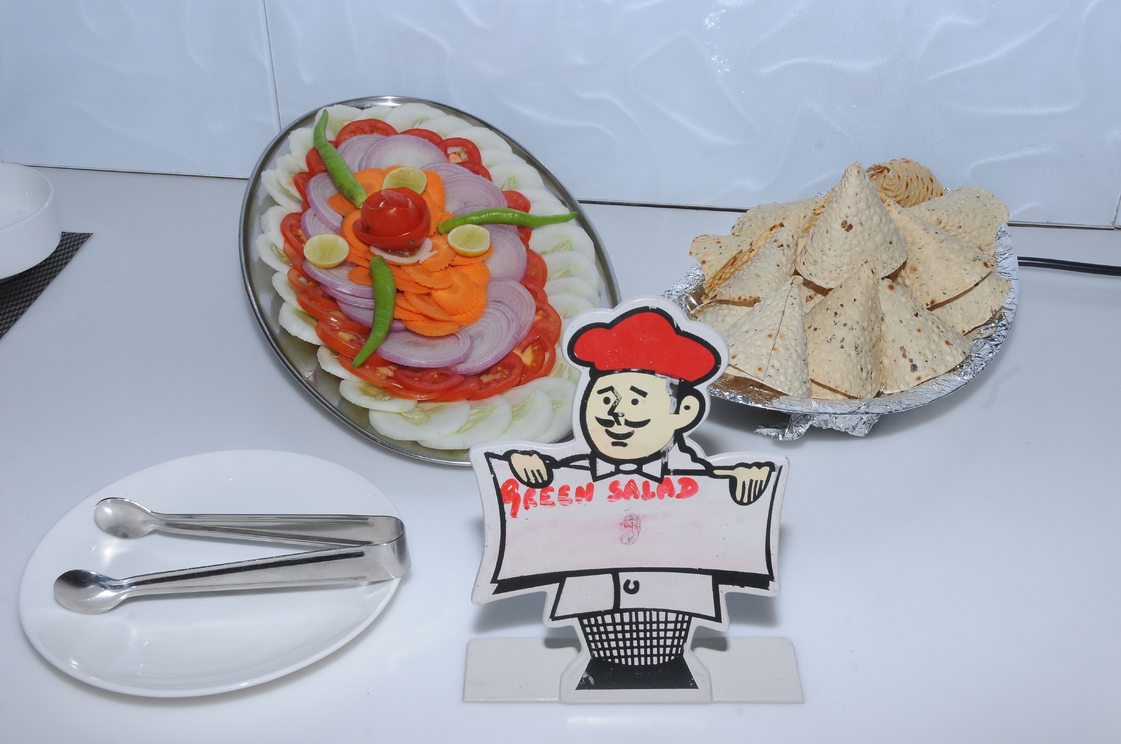 A plate of sliced vegetables, a plate of tortilla chips, and a sign with a chef illustration.