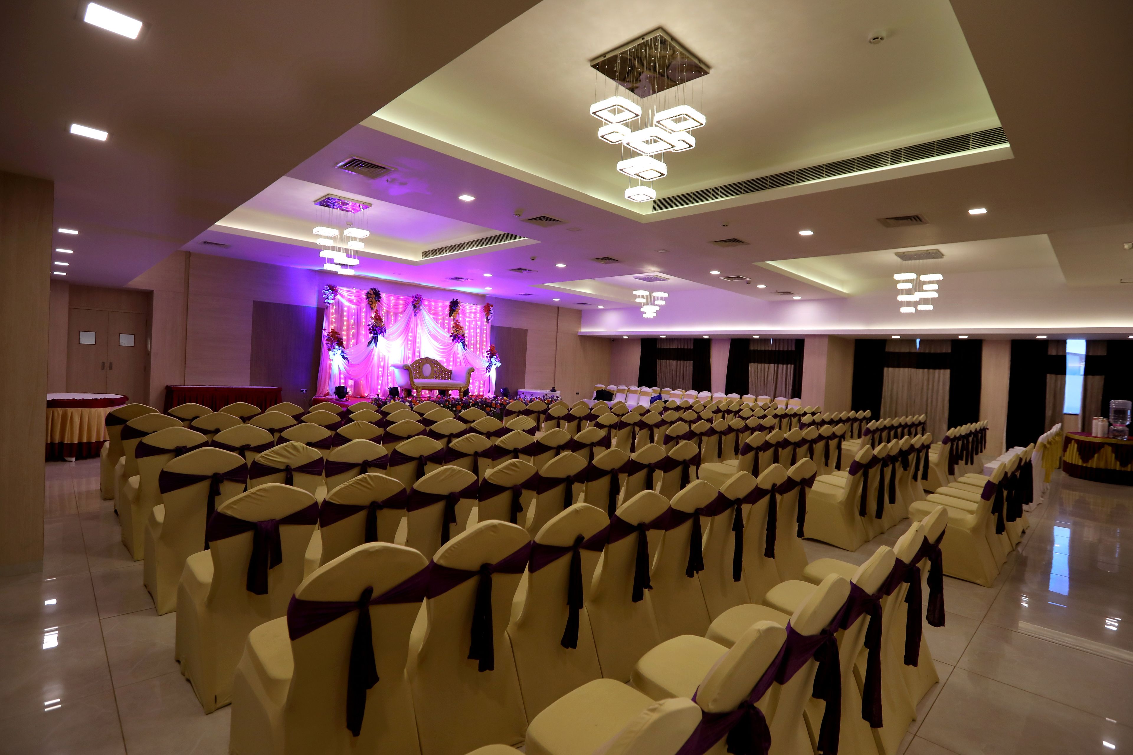 A banquet hall with rows of chairs, a stage with purple lighting, and modern ceiling fixtures.
