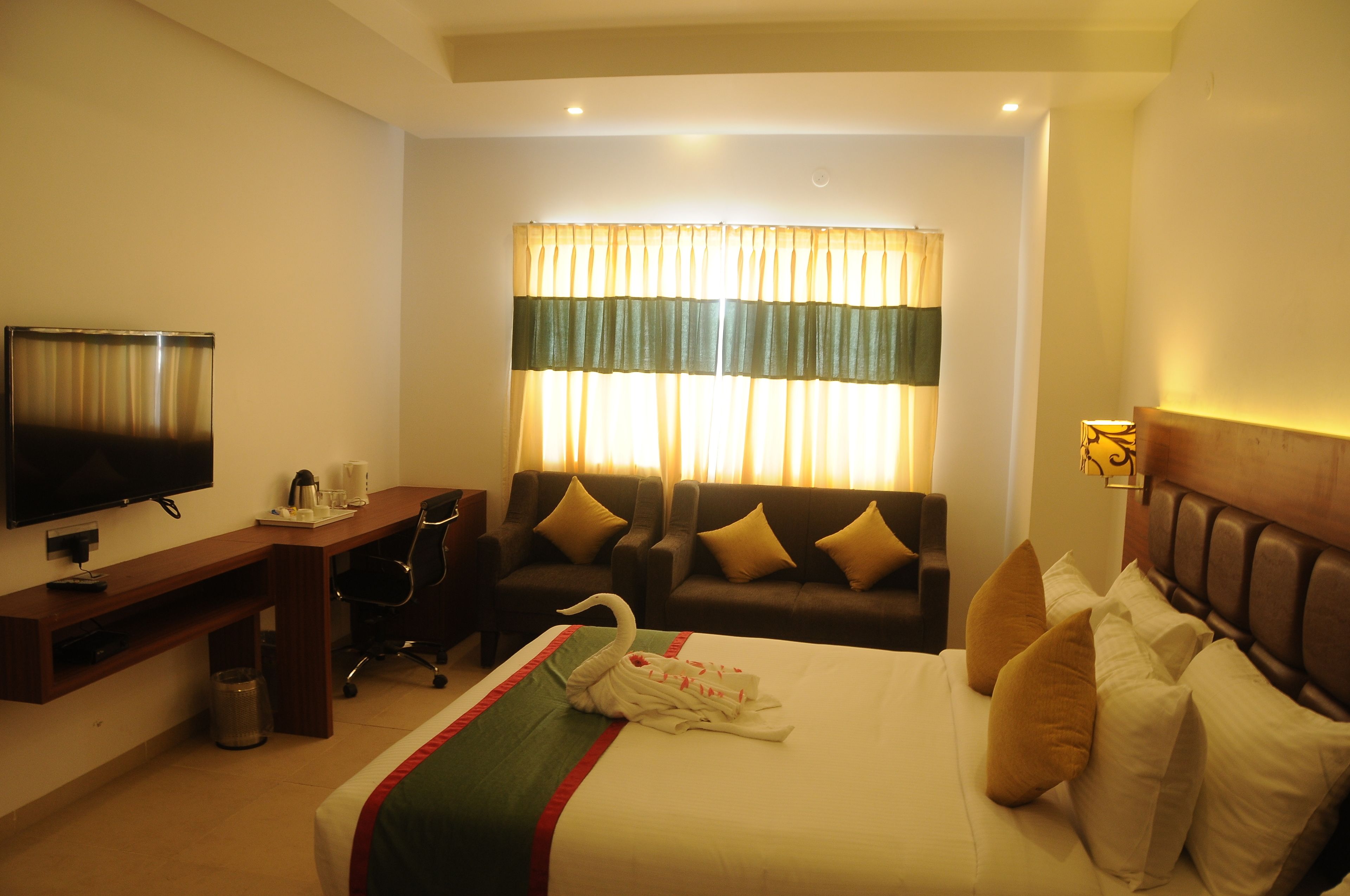 A hotel room with a bed, a TV, a desk, a chair, a sofa, and a window with curtains.
