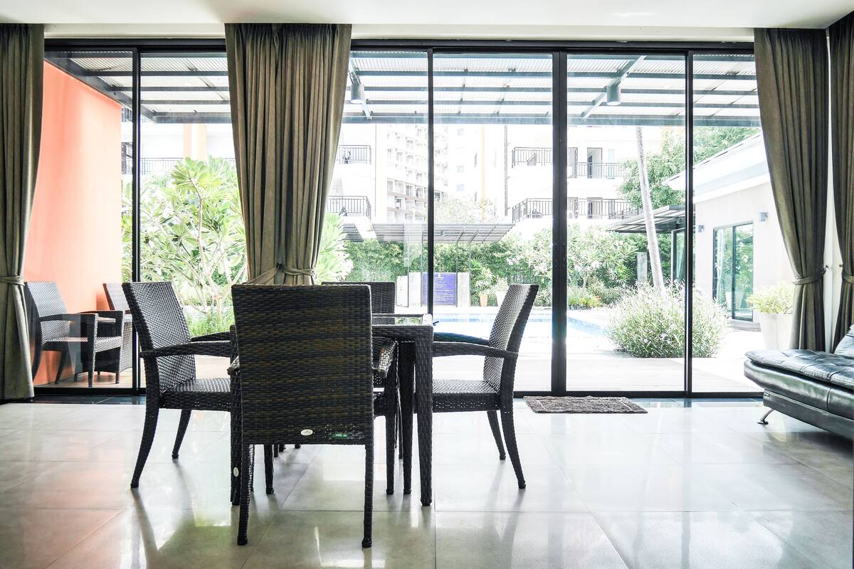 A dining area with wicker chairs and a table, large windows with curtains, and a view of a building and greenery outside.