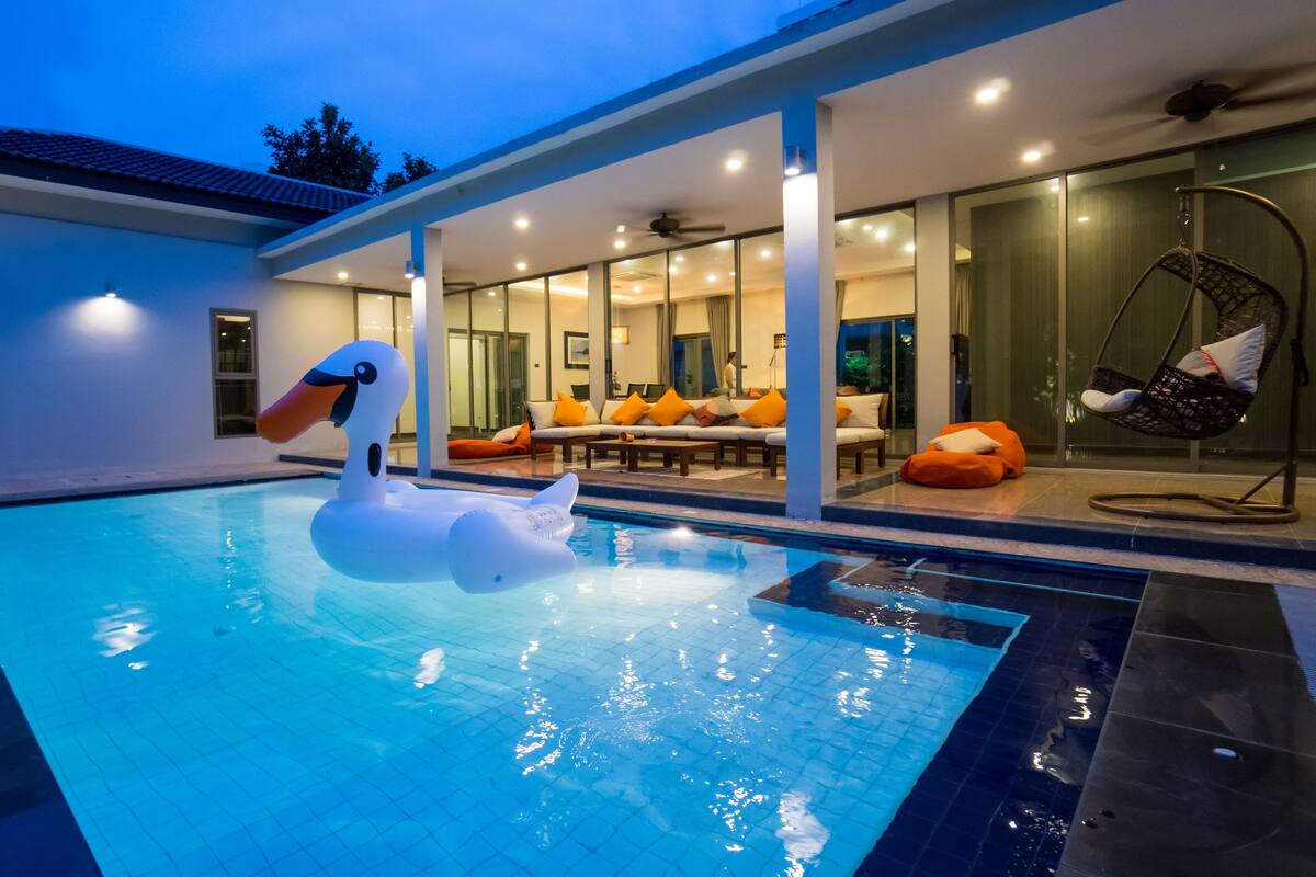 A swimming pool with a swan float, a covered outdoor seating area with orange cushions, and a hanging chair.