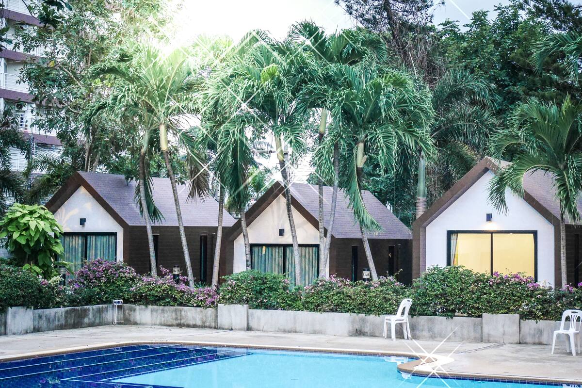 A pool area with a clear blue pool, surrounded by palm trees and a row of small, single-story buildings with sloped roofs.