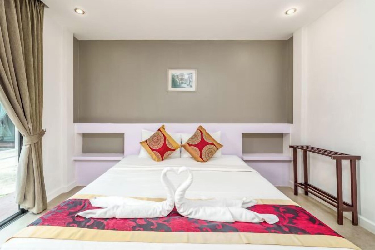 A hotel room with a large bed, two decorative pillows, a bedside table, and a window with curtains.