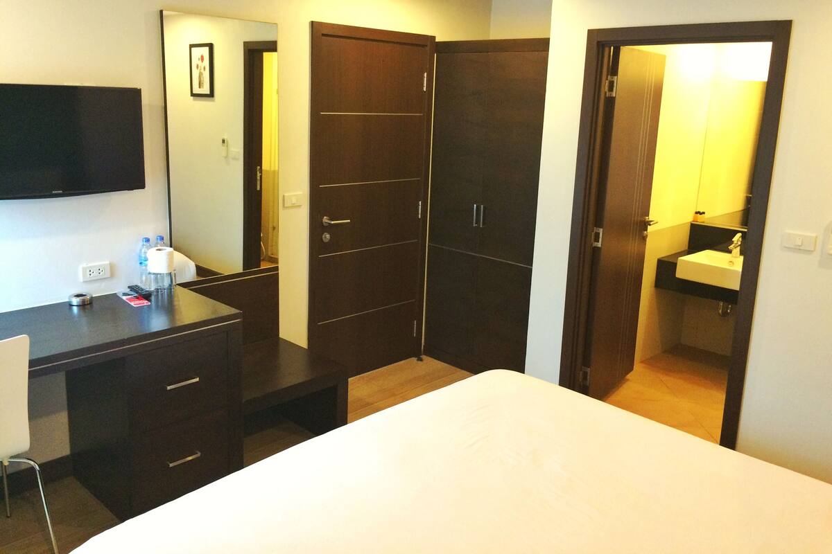 A hotel room with a bed, a desk with a chair, a wardrobe, a bathroom with a sink, and a flat-screen TV mounted on the wall.