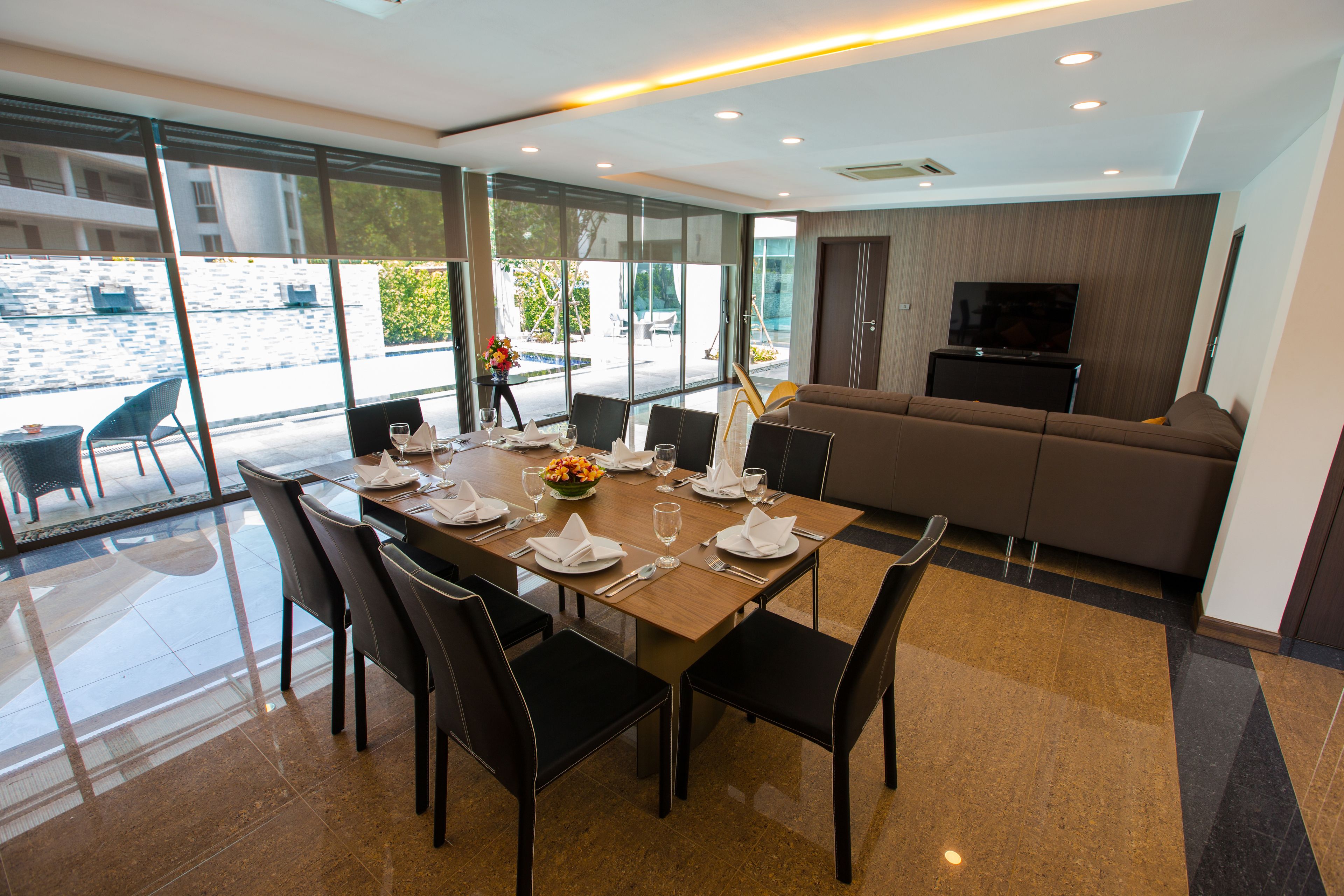 Un espace repas moderne avec une grande table dressée pour six personnes, entourée de chaises noires. On aperçoit un canapé et un téléviseur à l’arrière-plan.