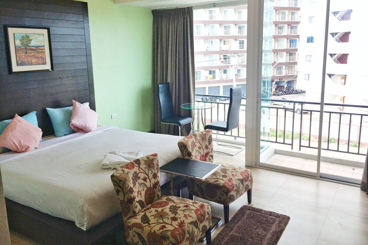 A hotel room with a large bed, floral-patterned armchairs, a small table, and a balcony with a view of a building.