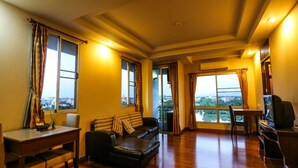 A living room with a sofa, a dining table, and a TV. There are large windows with curtains. - Madina Hotel (Rayong)