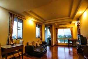 A living room with a sofa, a dining table, and a TV. There are large windows with curtains.