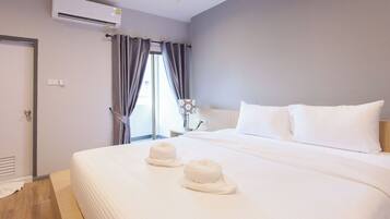 A hotel room with a large bed, white bedding, a window with curtains, an air conditioning unit, and a bedside table with a lamp.