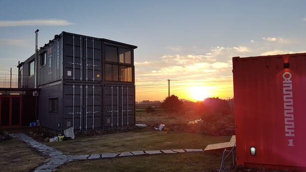 Un bâtiment moderne doté de grandes fenêtres et d’un toit plat, situé dans un espace verdoyant, avec un coucher de soleil en arrière-plan.