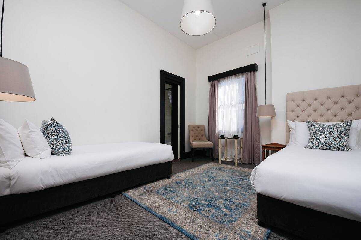 Une chambre d’hôtel avec deux lits simples, une petite table, une chaise et une grande fenêtre avec des rideaux.